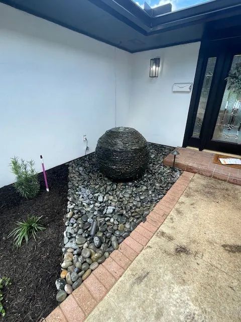 Front yard water fountain. Large, textured ball fountain on dark pebbles, bordered by brick and mulch.