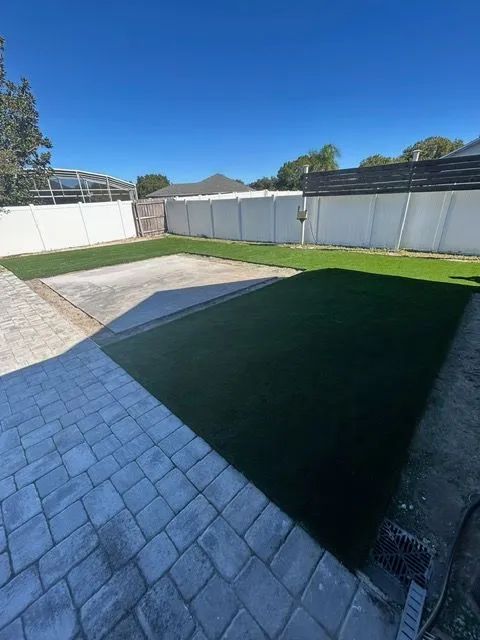 Backyard with gray pavers, concrete slab, and green artificial turf against white fence, under blue sky.