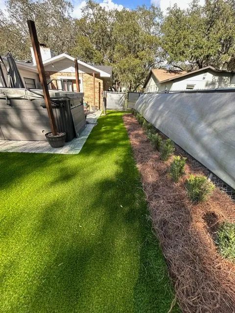 Backyard with green turf, a narrow bed with plants, and a light-colored wall.