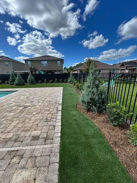 Backyard with patio, lawn, and landscaping. Blue sky with clouds. Houses with black fencing in the background.