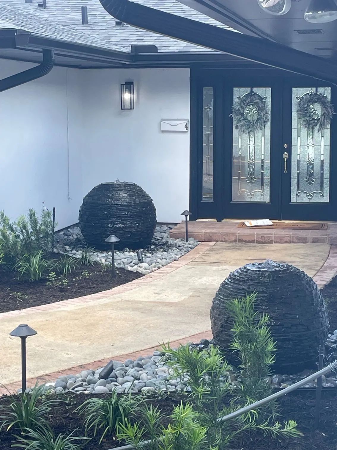 Black sphere fountains on either side of a walkway leading to a house with black double doors.