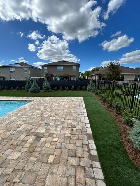 Backyard patio with pool, lawn, and bordering houses under a bright, cloudy sky.