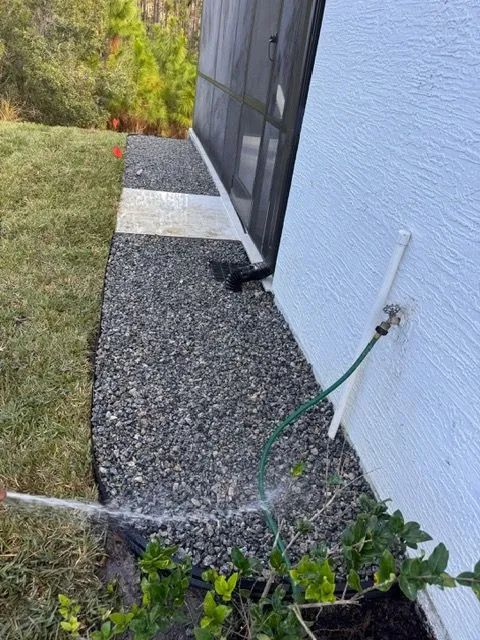Gravel walkway alongside a house being watered with a green hose, next to grass and bushes.
