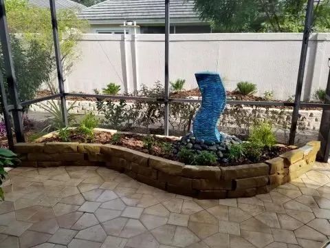 A decorative garden bed with a blue fountain, surrounded by stone pavers, shrubs, and a screened enclosure.