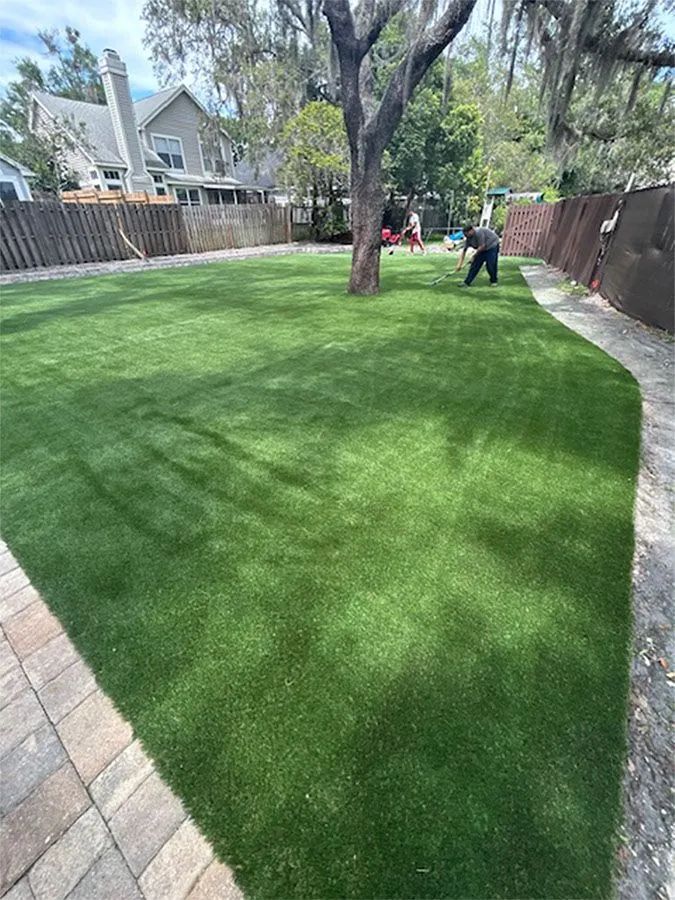 A backyard with green turf and a tree. A person works on the grass.