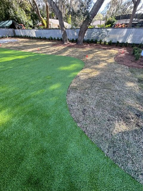 Half green artificial turf, half brown dead grass in a yard. Trees and a hedge in the background.