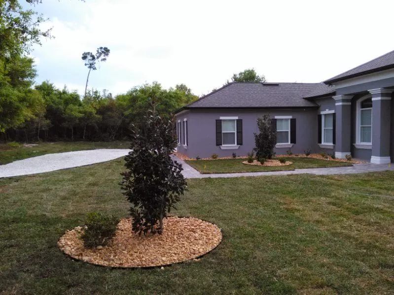 Gray house with dark shutters and roof. A tree and small bushes sit in a gravel bed on the front lawn.