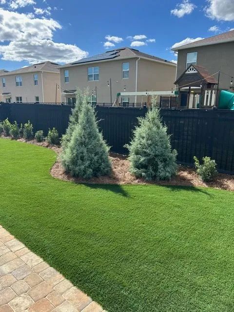 Backyard with green artificial grass, trees, a black fence, and houses on a sunny day.