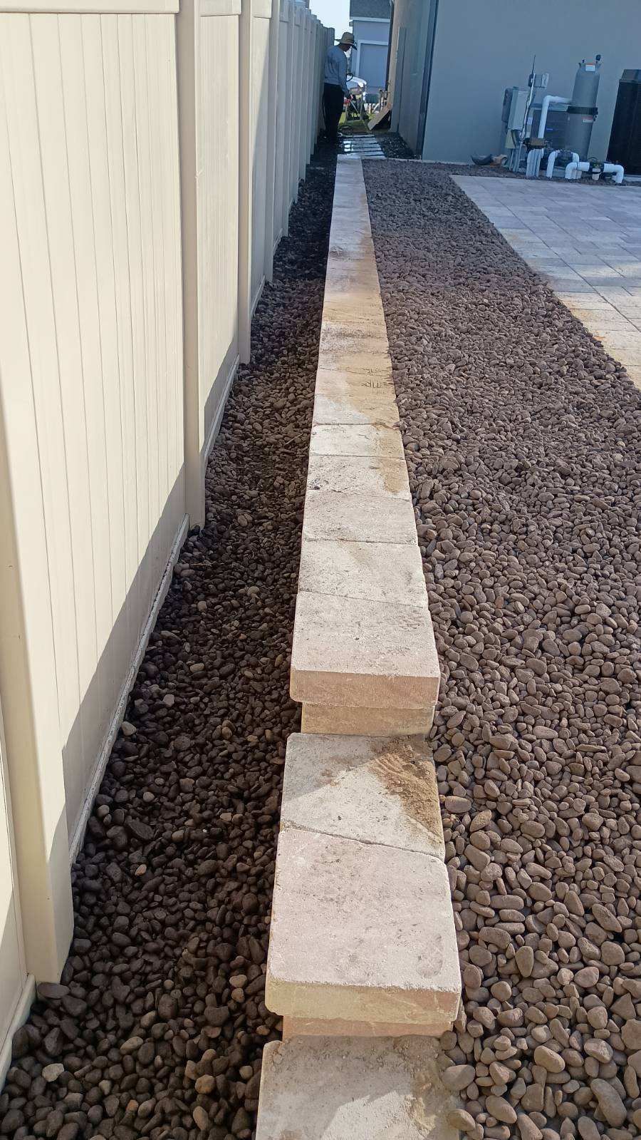 A long retaining wall of rectangular blocks next to a beige fence and dark gravel pathway.