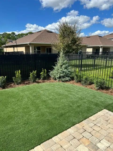 Green lawn with landscaped garden, black fence, and houses under blue sky.