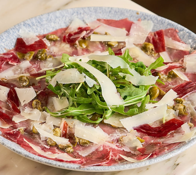 Salada de carne curada fatiada finamente com rúcula, queijo ralado, alcaparras e azeite de oliva em um prato.