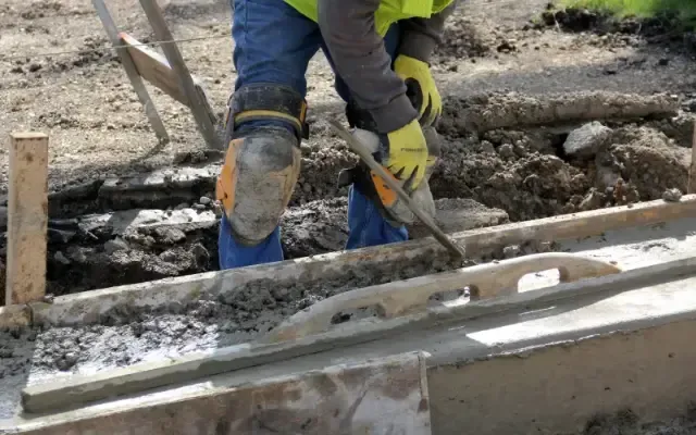 Construction worker smoothing concrete with a trowel, wearing gloves, knee pads, and safety vest.