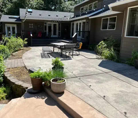 Backyard concrete patio with, brown house, and garden beds. Outdoor table and chairs.