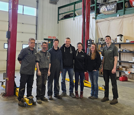 Seven people standing together in an auto repair shop, smiling in front of a car lift.