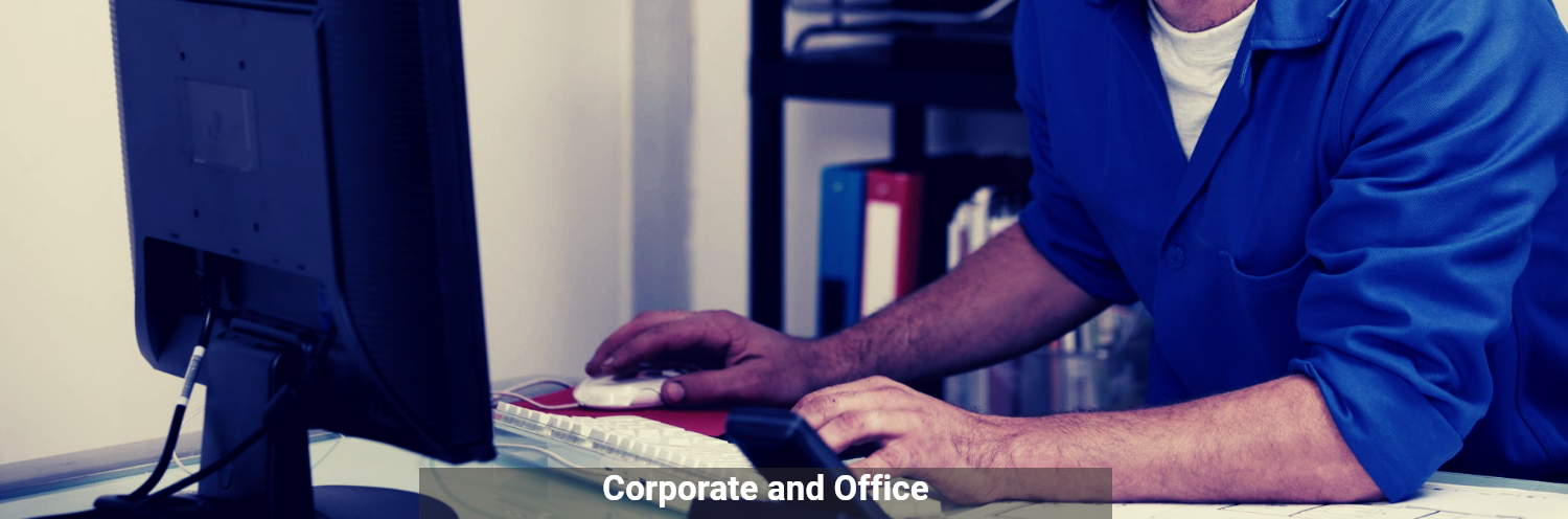 A person in a blue shirt works at a computer in an office setting with binders in the background.