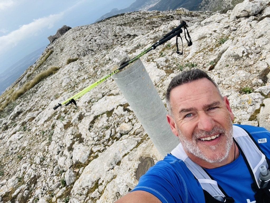 Un hombre con una camisa azul se está tomando una selfie en la cima de una montaña rocosa.