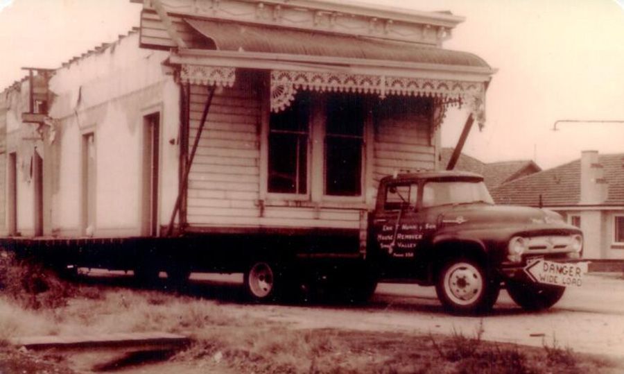 A black and white photo of a truck carrying a house on a trailer.