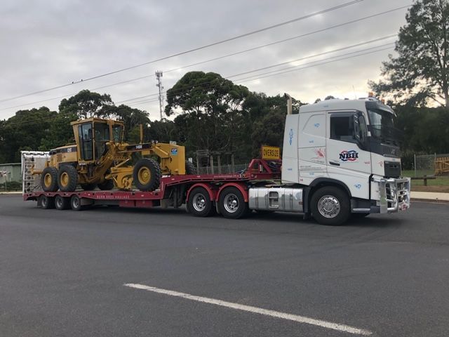 A white semi truck is carrying a yellow tractor on a trailer.