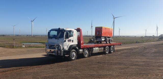 A tow truck is driving down a dirt road with windmills in the background.