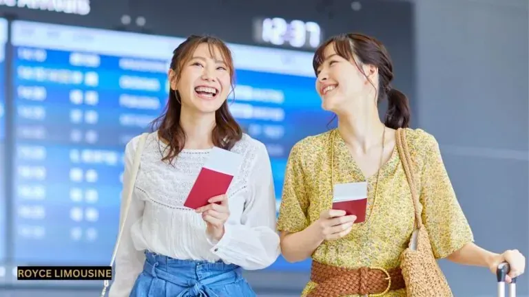 Two women smiling, holding passports, in an airport, near a departure board.