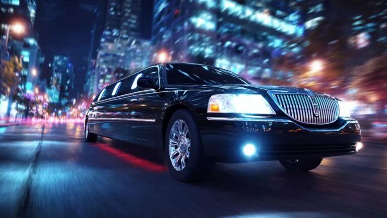 Black limousine driving quickly down a city street at night, headlights on.