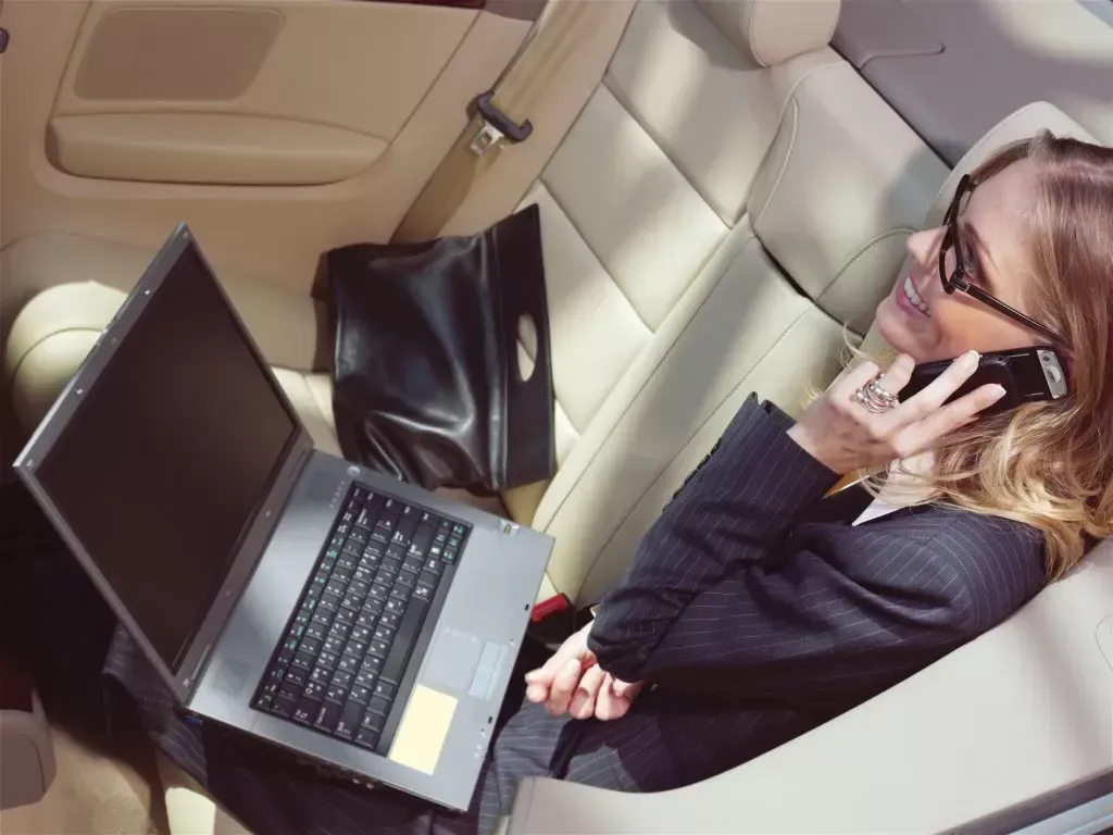 Woman in business suit, on phone, working on laptop in car back seat.