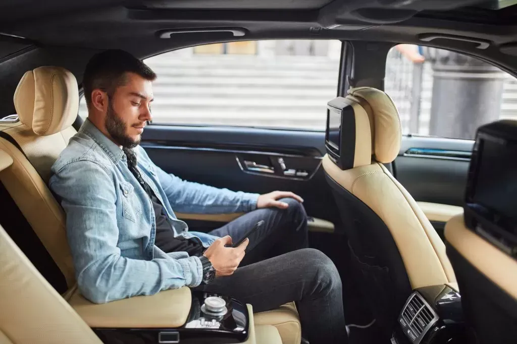 Man in back seat of car, looking at phone. Beige leather seats, dark interior, daytime.