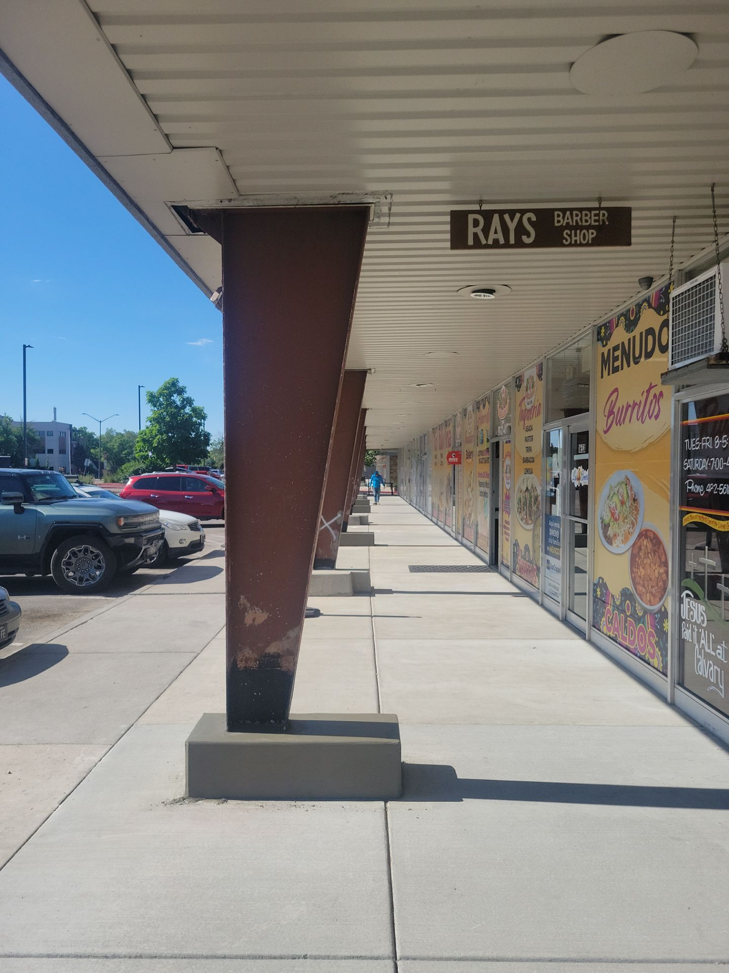 A sunny outdoor storefront, with shops and a sidewalk lined with brown support beams.