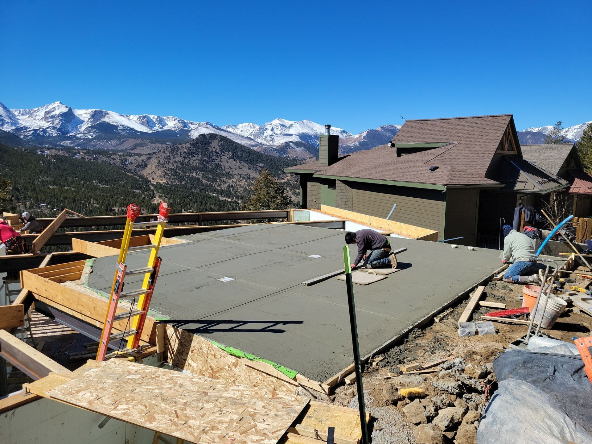Construction site with workers pouring concrete slab for a deck, mountains in background, sunny day.