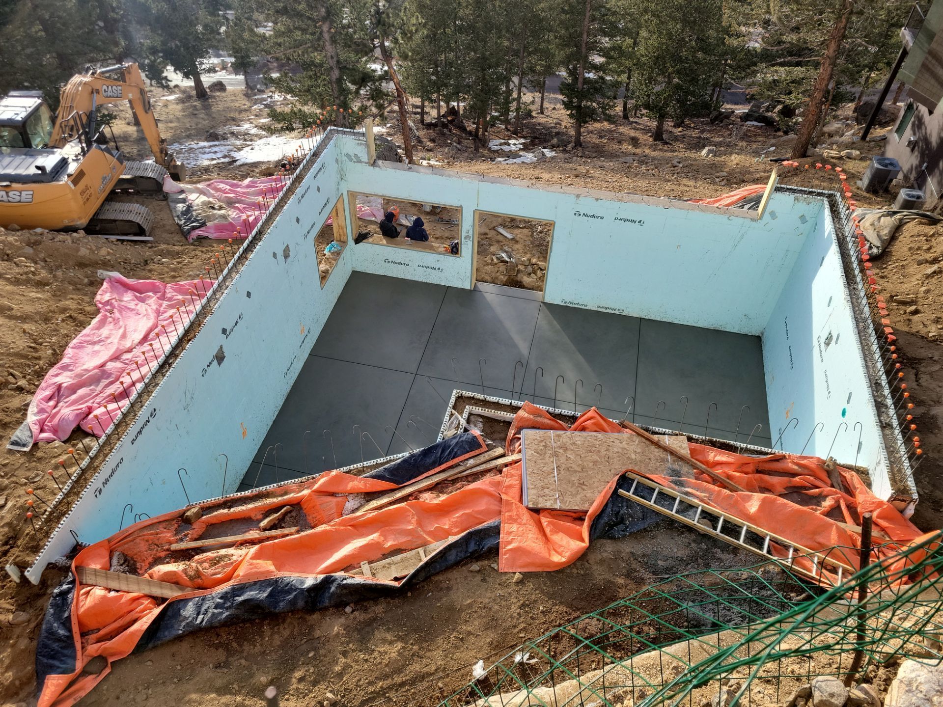Foundation walls with blue insulation, dirt surrounding. Construction site with excavator visible.