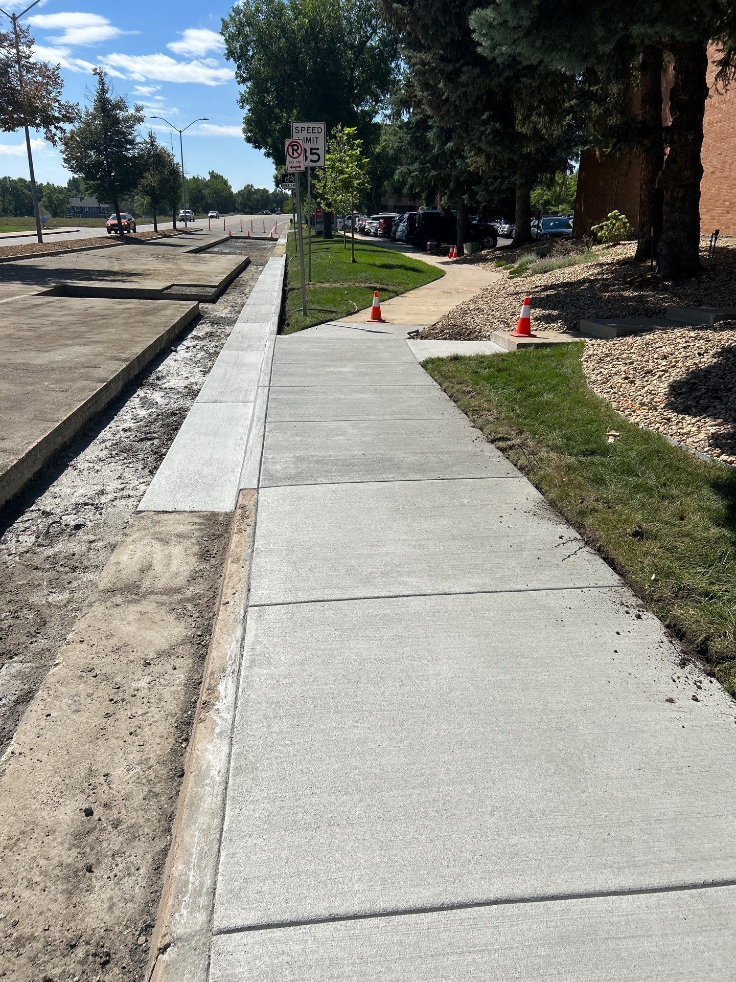 Newly constructed concrete sidewalk next to a curb and grass; street and buildings in background.