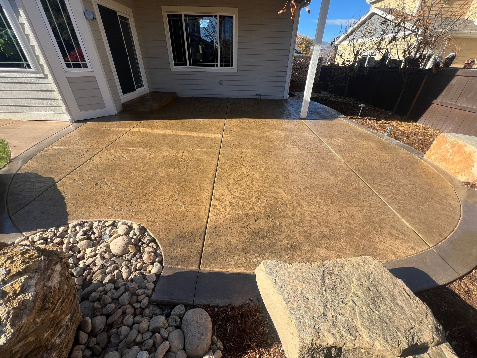 Concrete patio with curved edges, stained brown, next to a rock garden and house.