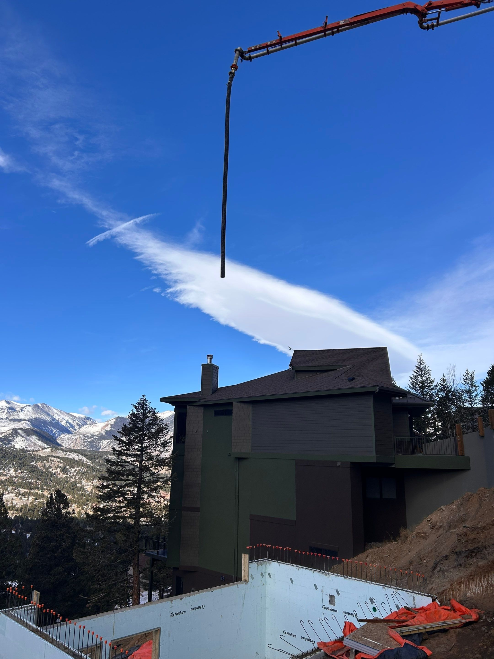 Construction site with a concrete pump arm over a dark green house and a partially built foundation, mountain backdrop.