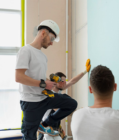 Construction workers installing drywall; one drilling, others steadying. Brightly lit interior.