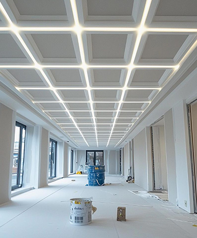 A long, white room under construction. Square ceiling lights, windows on the left, blue drum in center.