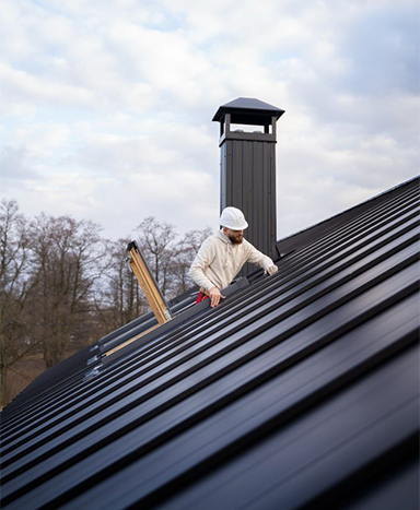Person on a black metal roof near a chimney, wearing a hard hat, working.