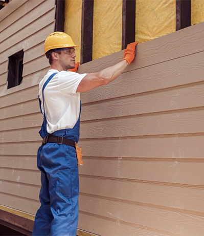 Construction worker installing siding on a building. He wears a hard hat and overalls, holding the siding in place.