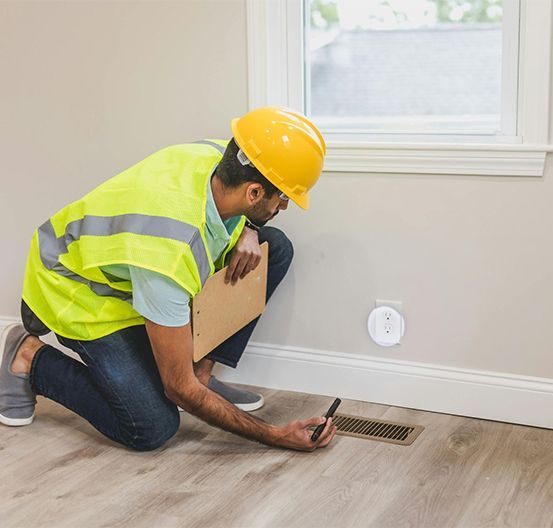 Inspector in hard hat and vest examines a floor vent with clipboard and pen in a room with a window.