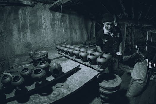Man arranging pottery jars in a dimly lit workshop. Jars sit on a table, another person is nearby.