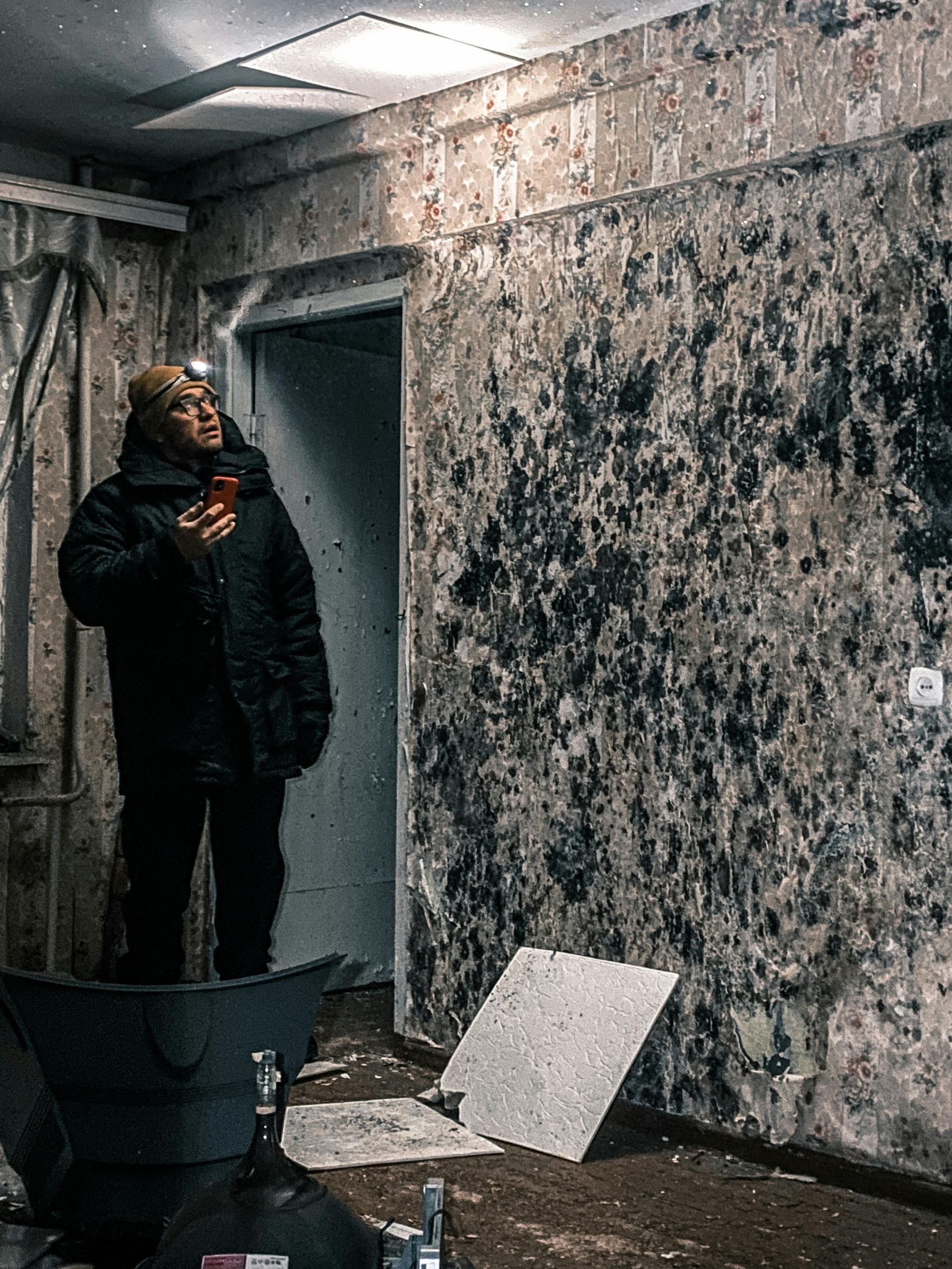 Man in dark coat with headlamp examines moldy room, holding phone.