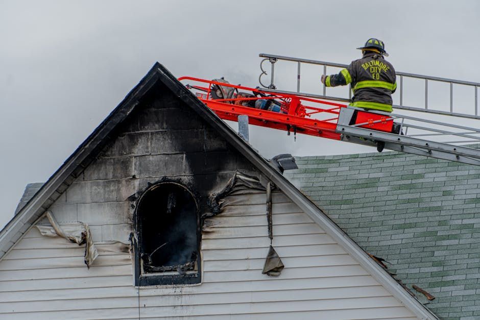 Firefighter on a ladder at a house fire, smoke billowing from a damaged window, grey sky.