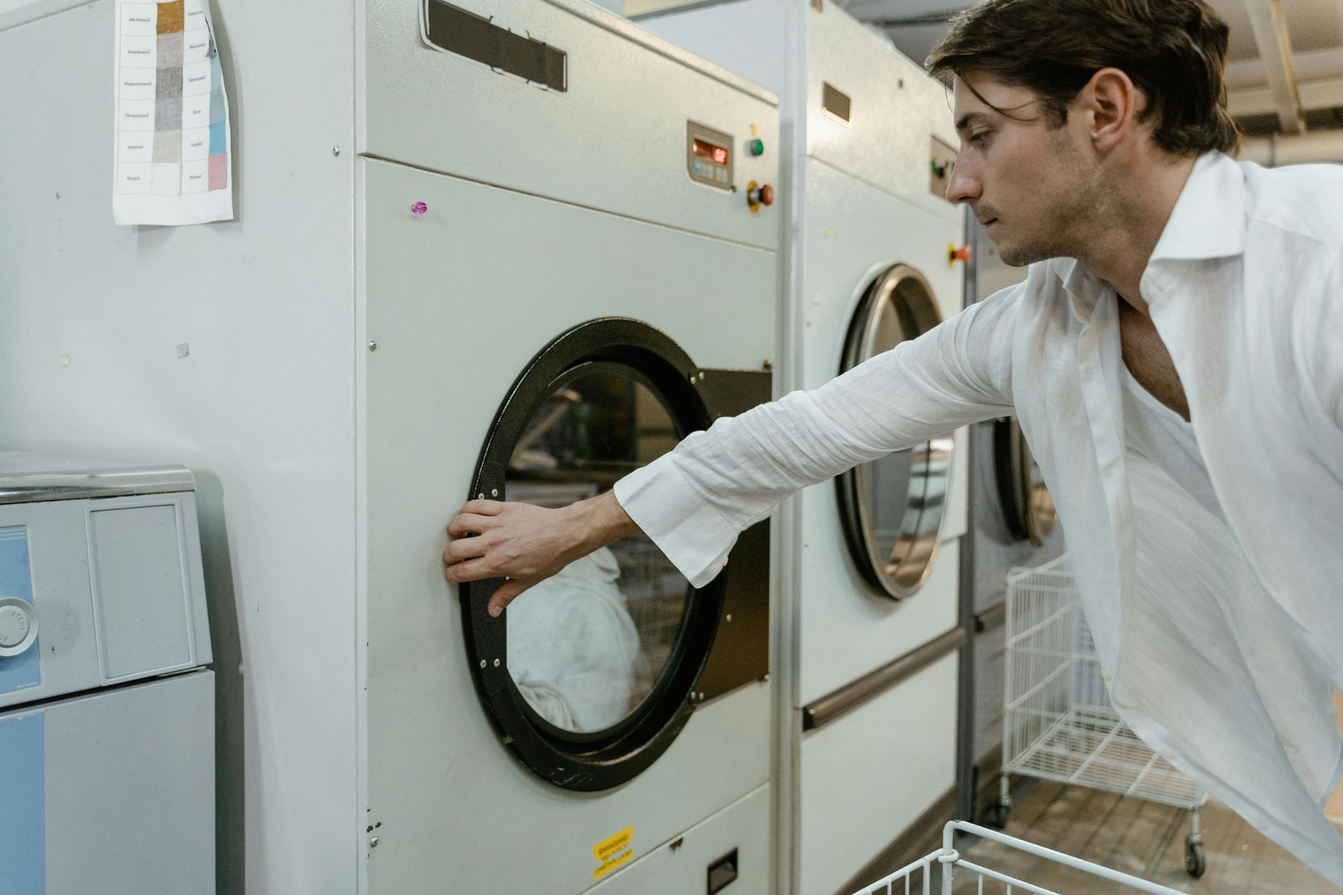 Man in white shirt opening a dryer door in a laundromat.