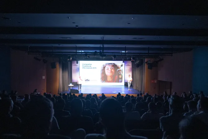 Público assistindo a uma apresentação em uma tela grande em um auditório escuro.