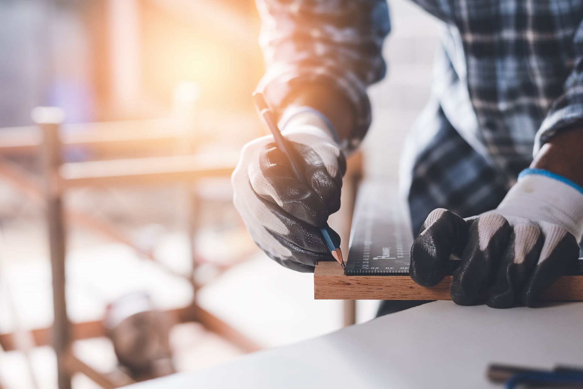 A person wearing work gloves marks a measurement on a wooden board with a pencil and a metal ruler.