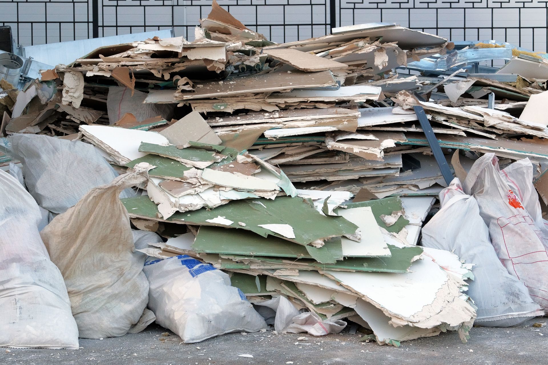 A large pile of construction debris, including pieces of drywall and plastic waste bags, sits against a metal fence.