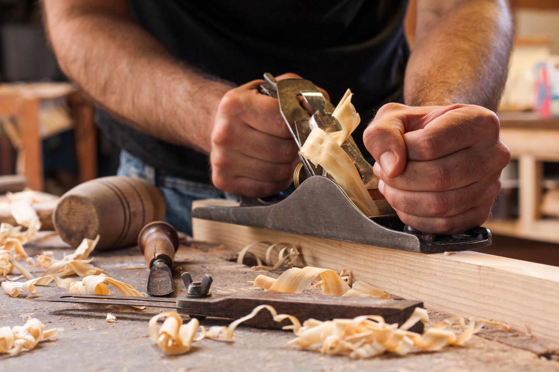 A person using a hand plane to smooth a wooden board on a workbench covered in wood shavings.