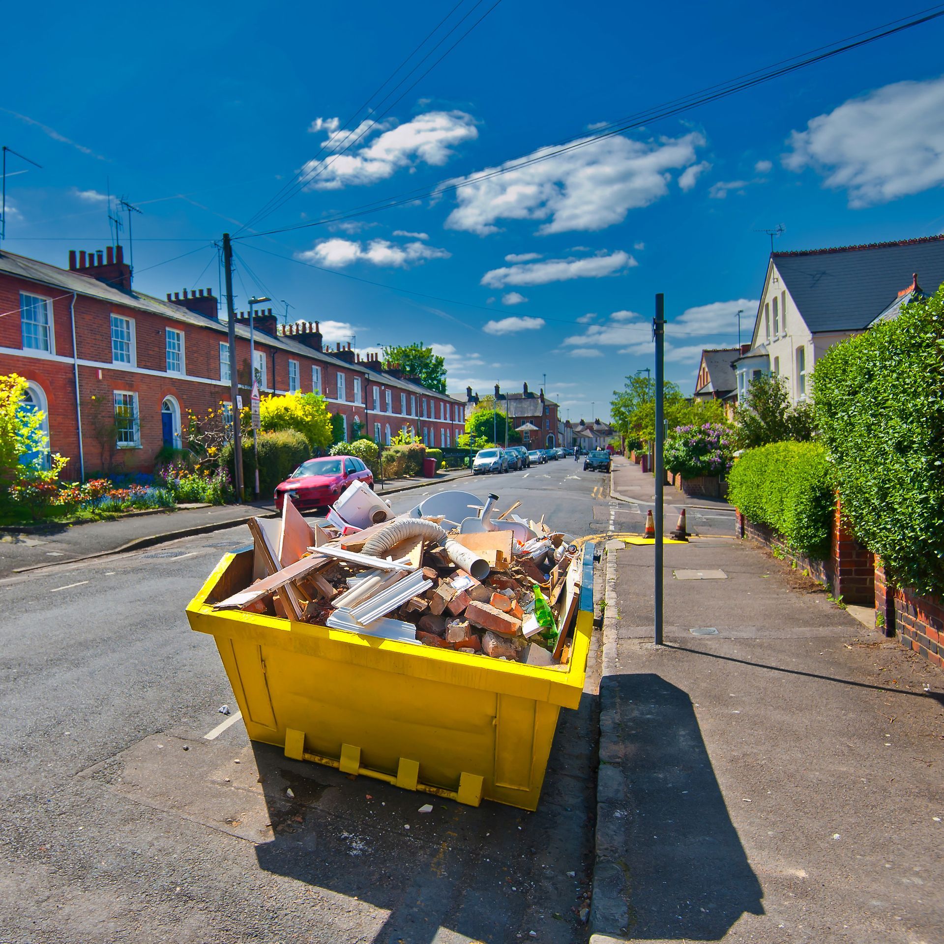 A bright yellow skip filled with construction debris sits on the side of a residential street under a blue sky.