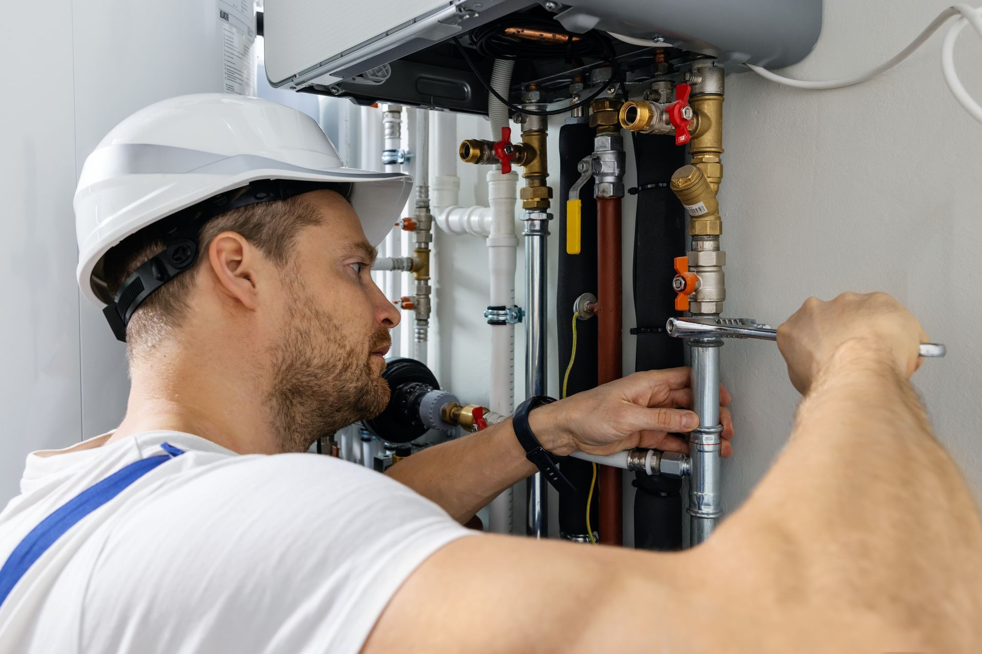 A technician in a hard hat repairs pipes with a wrench near a heating system.