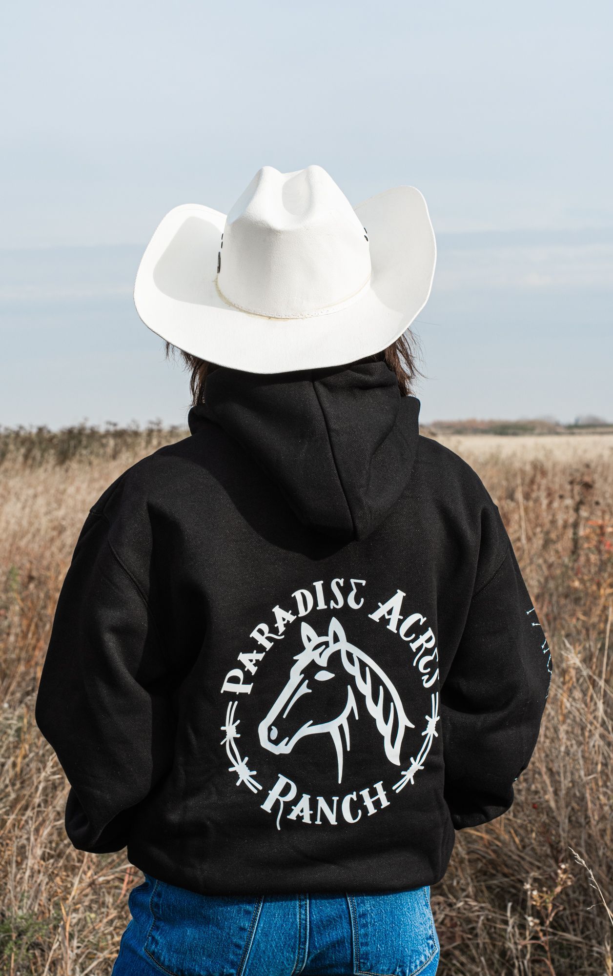 A woman wearing a cowboy hat and a black hoodie is standing in a field.