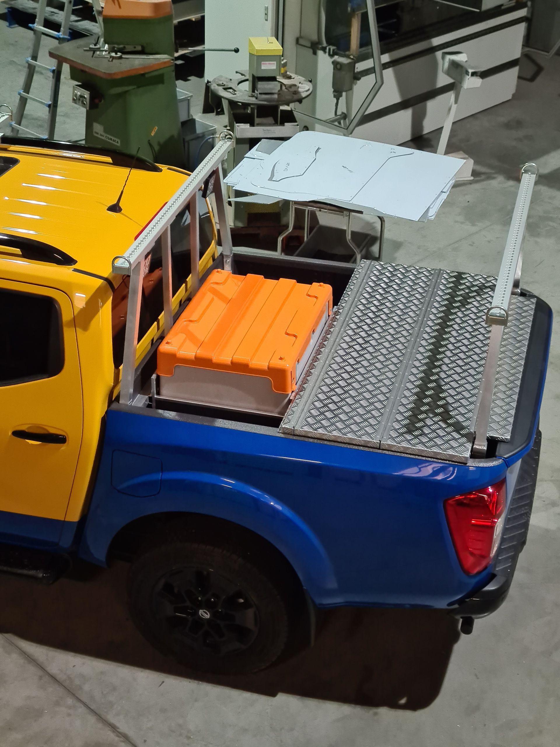 Blaue und gelbe Ladefläche eines Pickups mit orangefarbenen Containern und grauen Metallplatten, im Innenbereich geparkt.